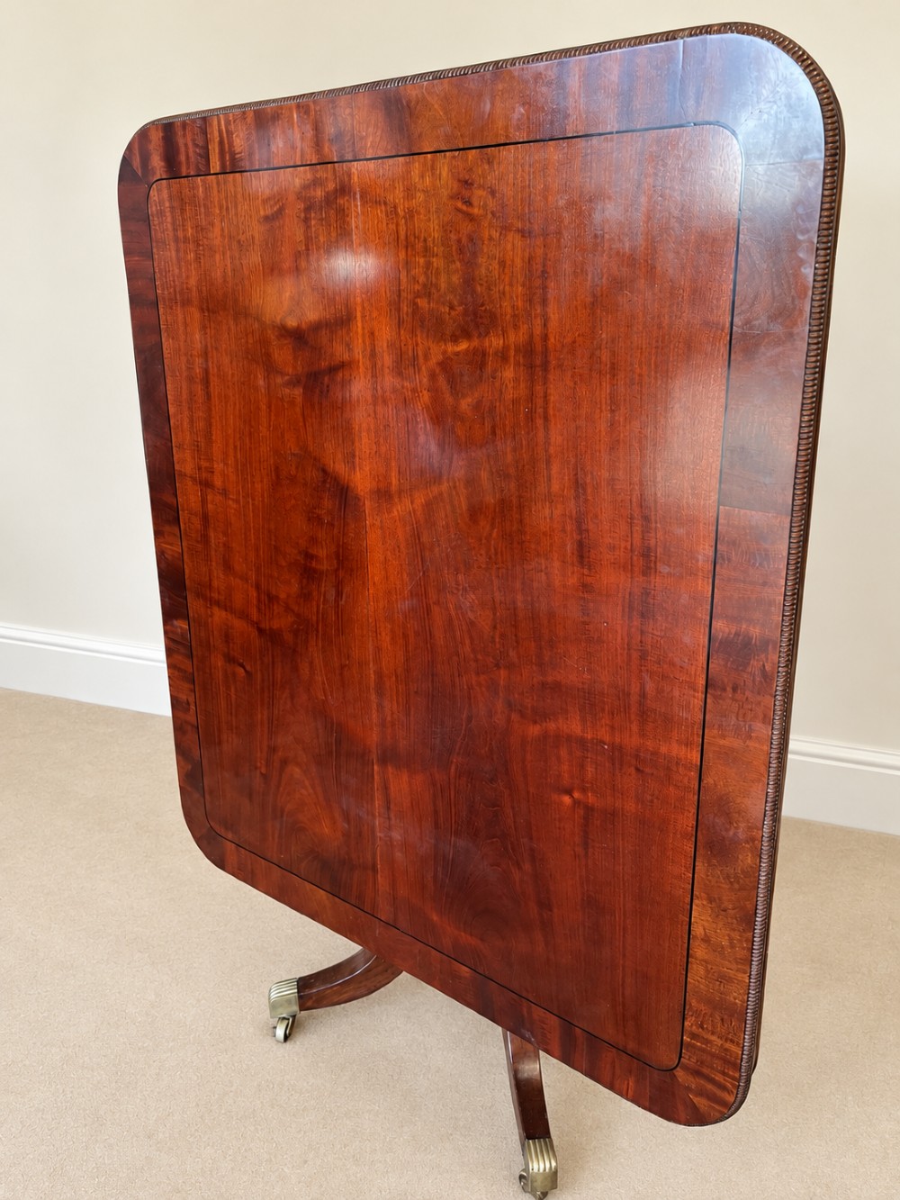 A Regency period mahogany dining table featuring a square top with rounded corners, supported by a central pedestal and four splayed legs with brass castors. View IX