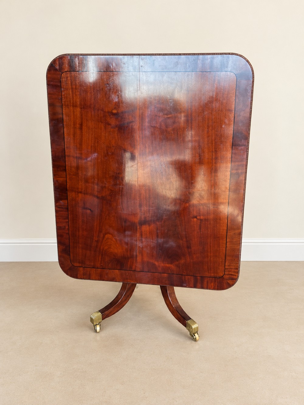 A Regency period mahogany dining table featuring a square top with rounded corners, supported by a central pedestal and four splayed legs with brass castors. View XI