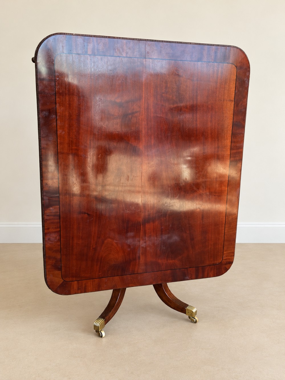 A Regency period mahogany dining table featuring a square top with rounded corners, supported by a central pedestal and four splayed legs with brass castors. View XII