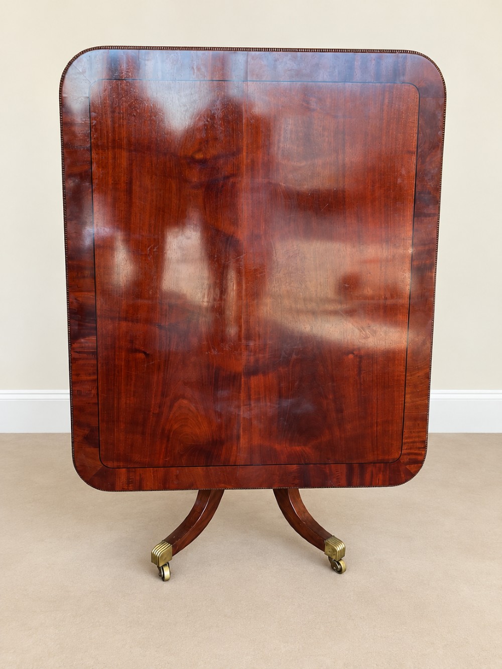A Regency period mahogany dining table featuring a square top with rounded corners, supported by a central pedestal and four splayed legs with brass castors. View XIII