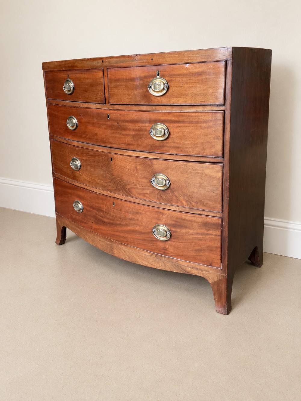 A nineteenth-century mahogany bowfront chest of drawers featuring two short drawers above three long graduated drawers with oval brass handles and splayed bracket feet. View V