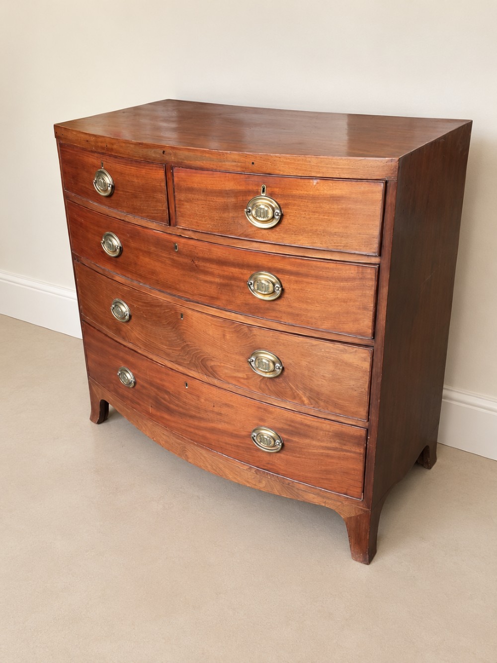 A nineteenth-century mahogany bowfront chest of drawers featuring two short drawers above three long graduated drawers with oval brass handles and splayed bracket feet. View VIII