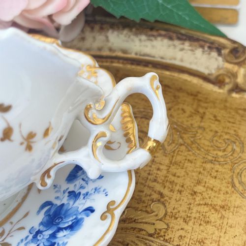 fig 1. An antique Regency period white porcelain teacup and saucer featuring hand-painted blue floral motifs and gilded ornate rim embellishments.