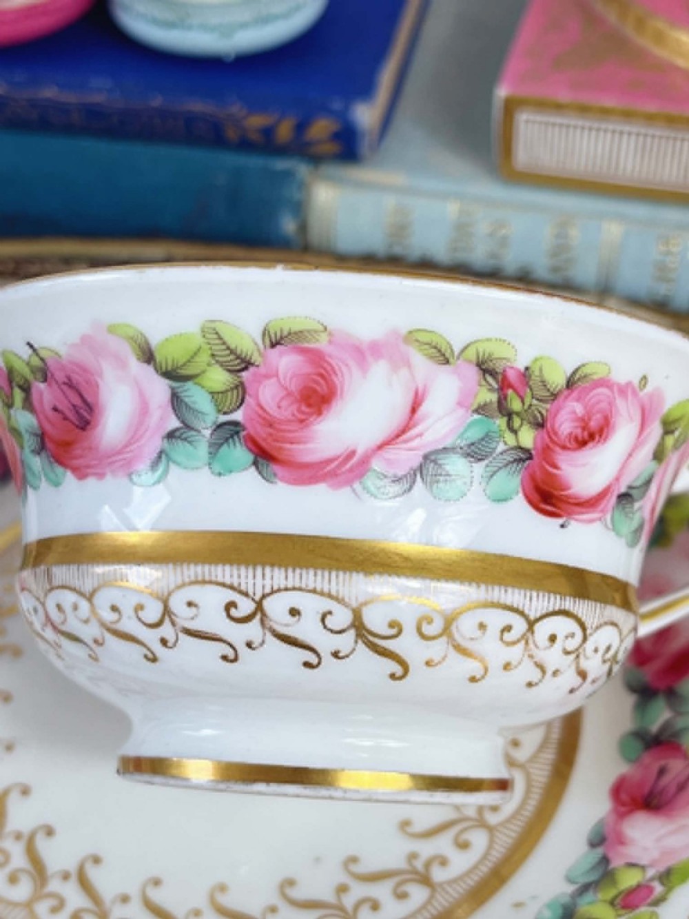 An antique porcelain teacup and matching saucer featuring hand-painted pink cabbage roses, green leaves, and gold scroll decorations. View X