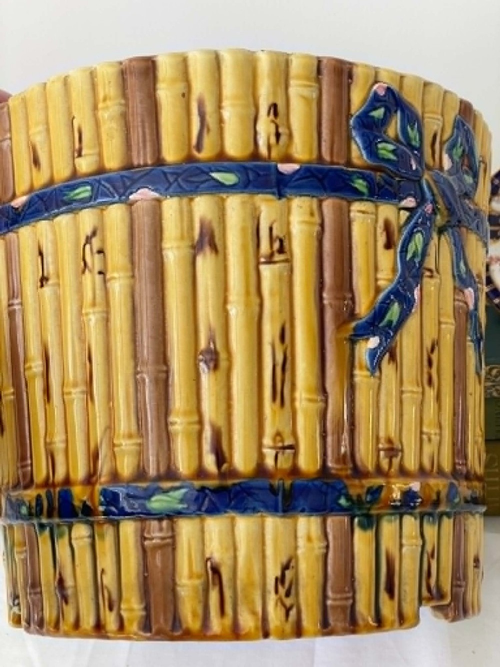 A cylindrical Victorian majolica jardiniere decorated with a moulded bamboo cane pattern in yellow and brown tones, wrapped with a blue ribbon tied in a bow. View IV