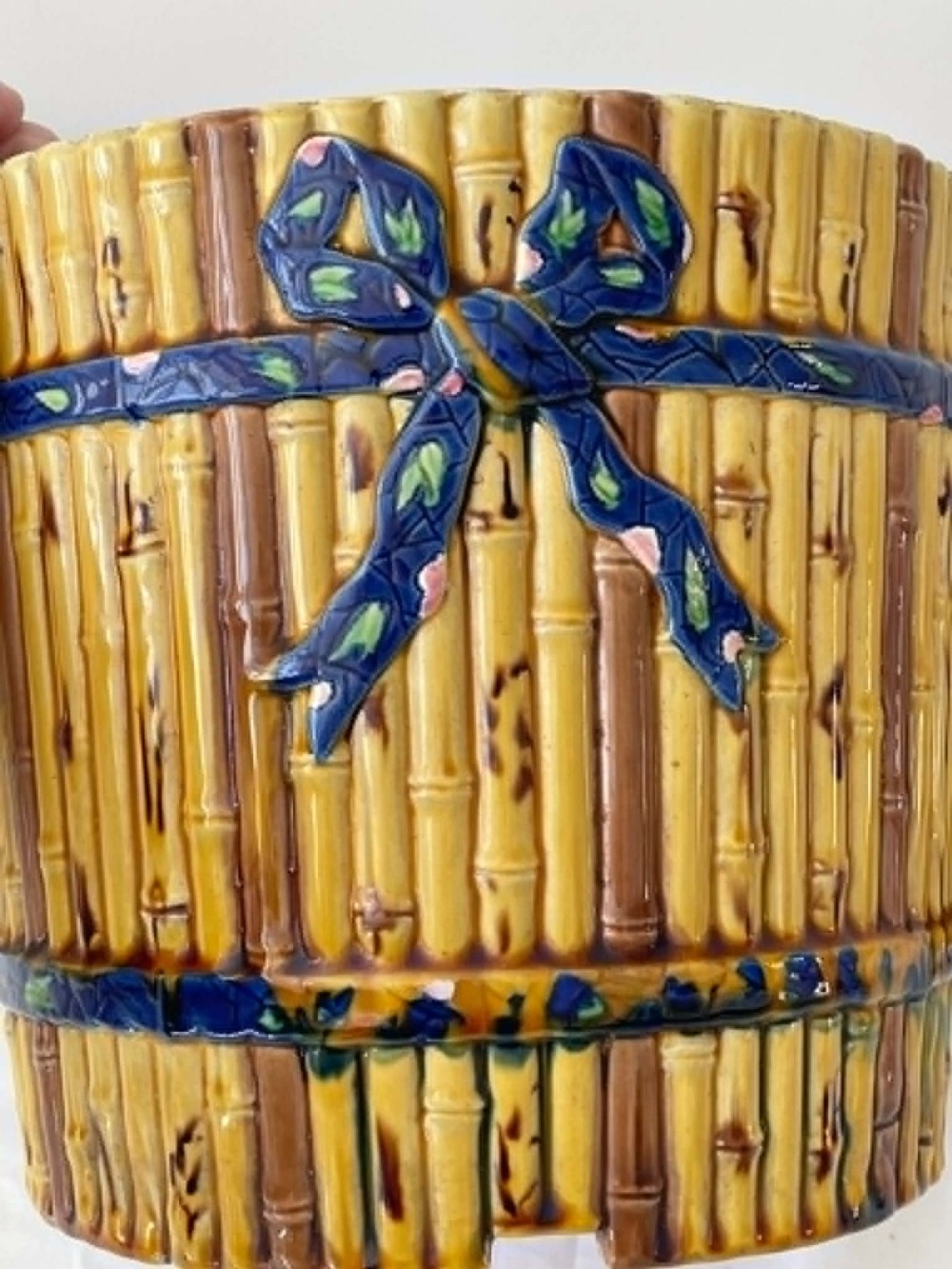 A cylindrical Victorian majolica jardiniere decorated with a moulded bamboo cane pattern in yellow and brown tones, wrapped with a blue ribbon tied in a bow. View V