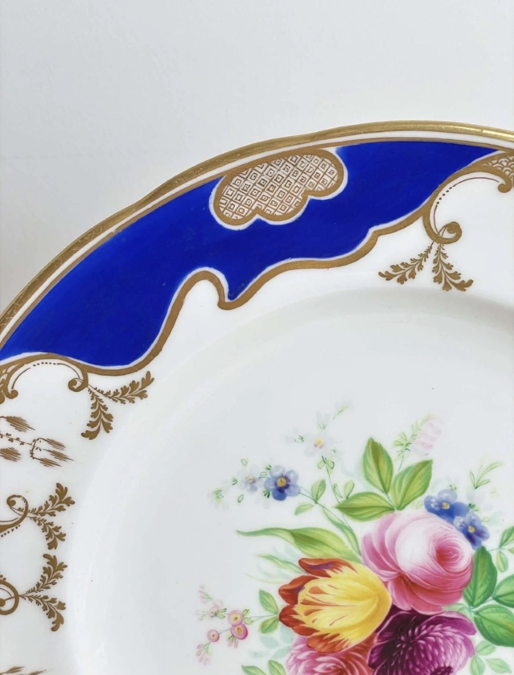 A circular porcelain dessert plate featuring a central hand-painted floral bouquet surrounded by a cobalt blue rim with gilded scrollwork. View VI