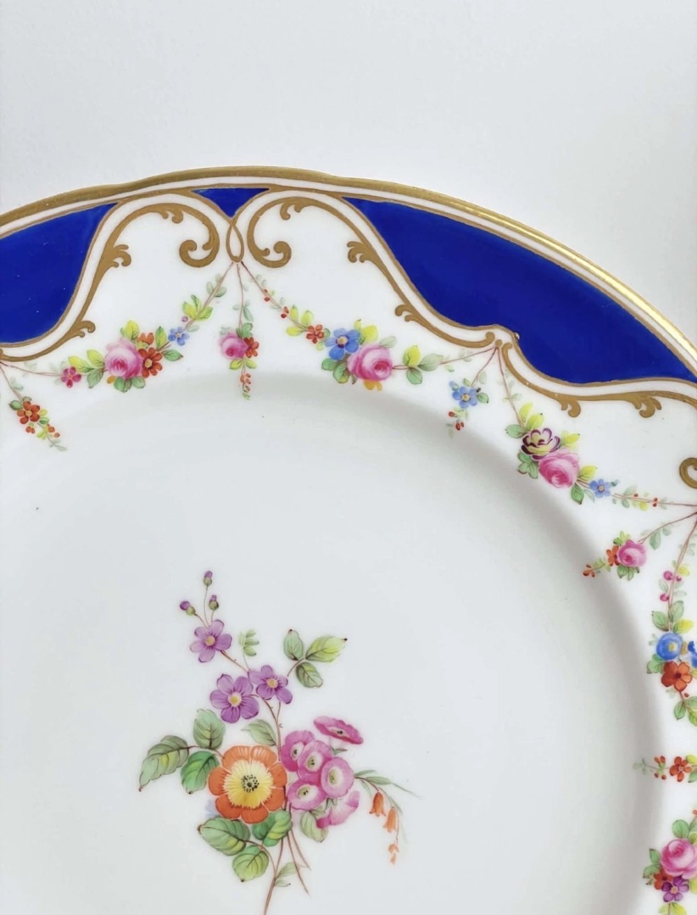 An antique circular porcelain dessert plate featuring a hand-painted central floral spray, delicate floral swags, and a scalloped cobalt blue and gilded rim. View V