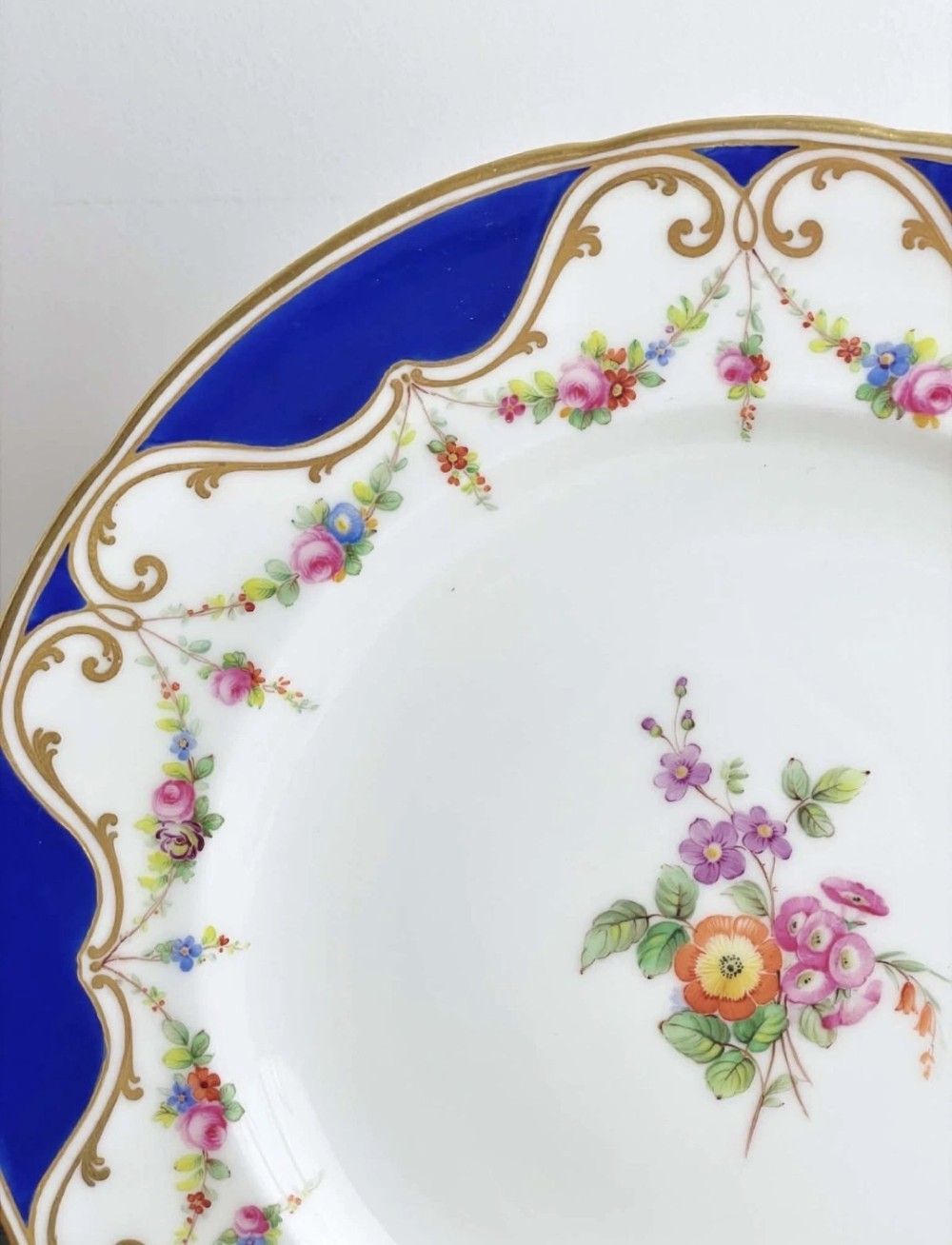 An antique circular porcelain dessert plate featuring a hand-painted central floral spray, delicate floral swags, and a scalloped cobalt blue and gilded rim. View VI