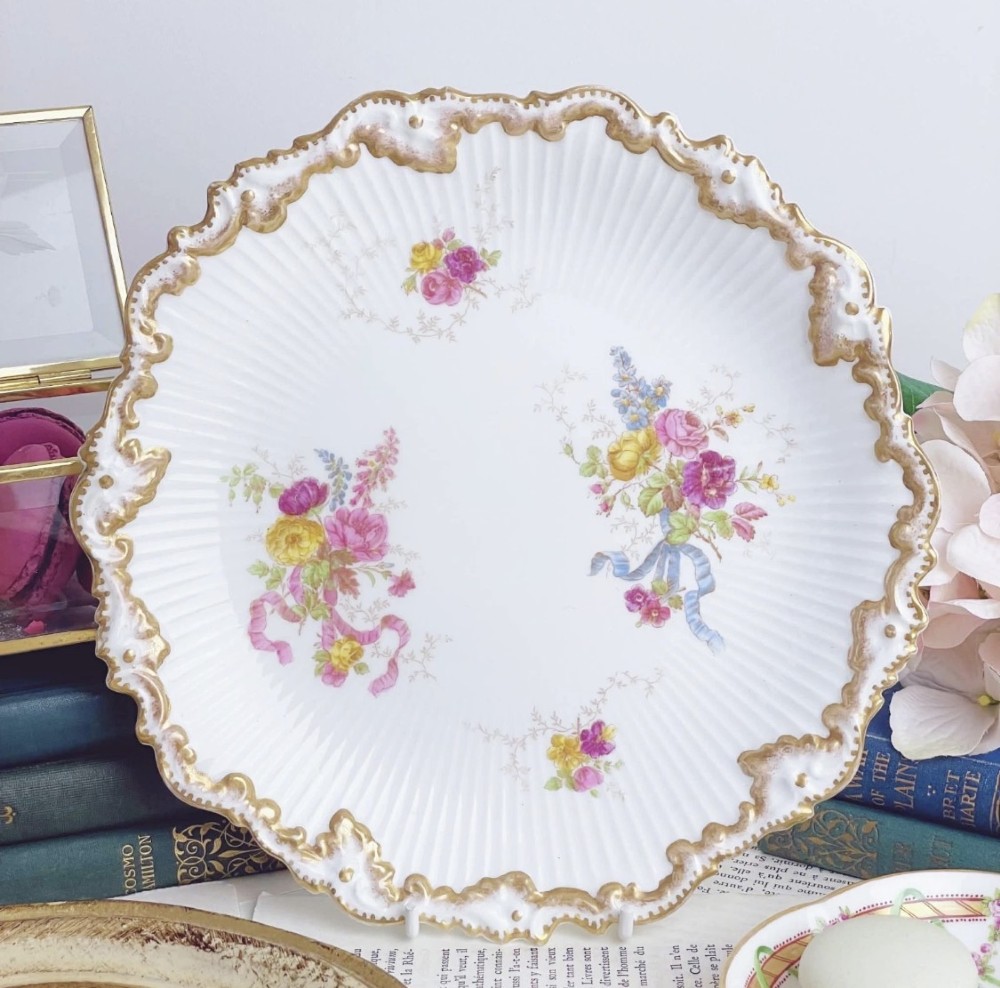 A white circular porcelain plate featuring a gilded scalloped rim, fluted texture, and hand-painted floral sprays adorned with pink and blue ribbons.