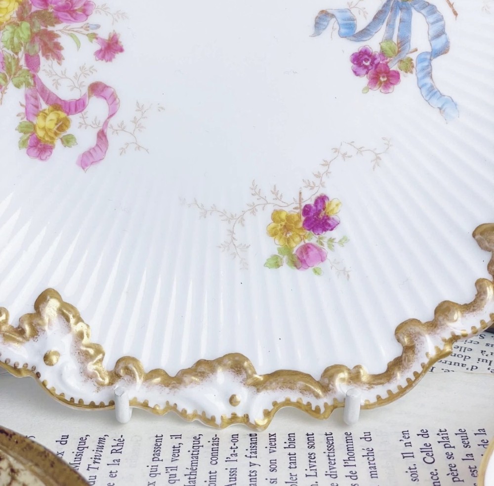 A white circular porcelain plate featuring a gilded scalloped rim, fluted texture, and hand-painted floral sprays adorned with pink and blue ribbons. View II
