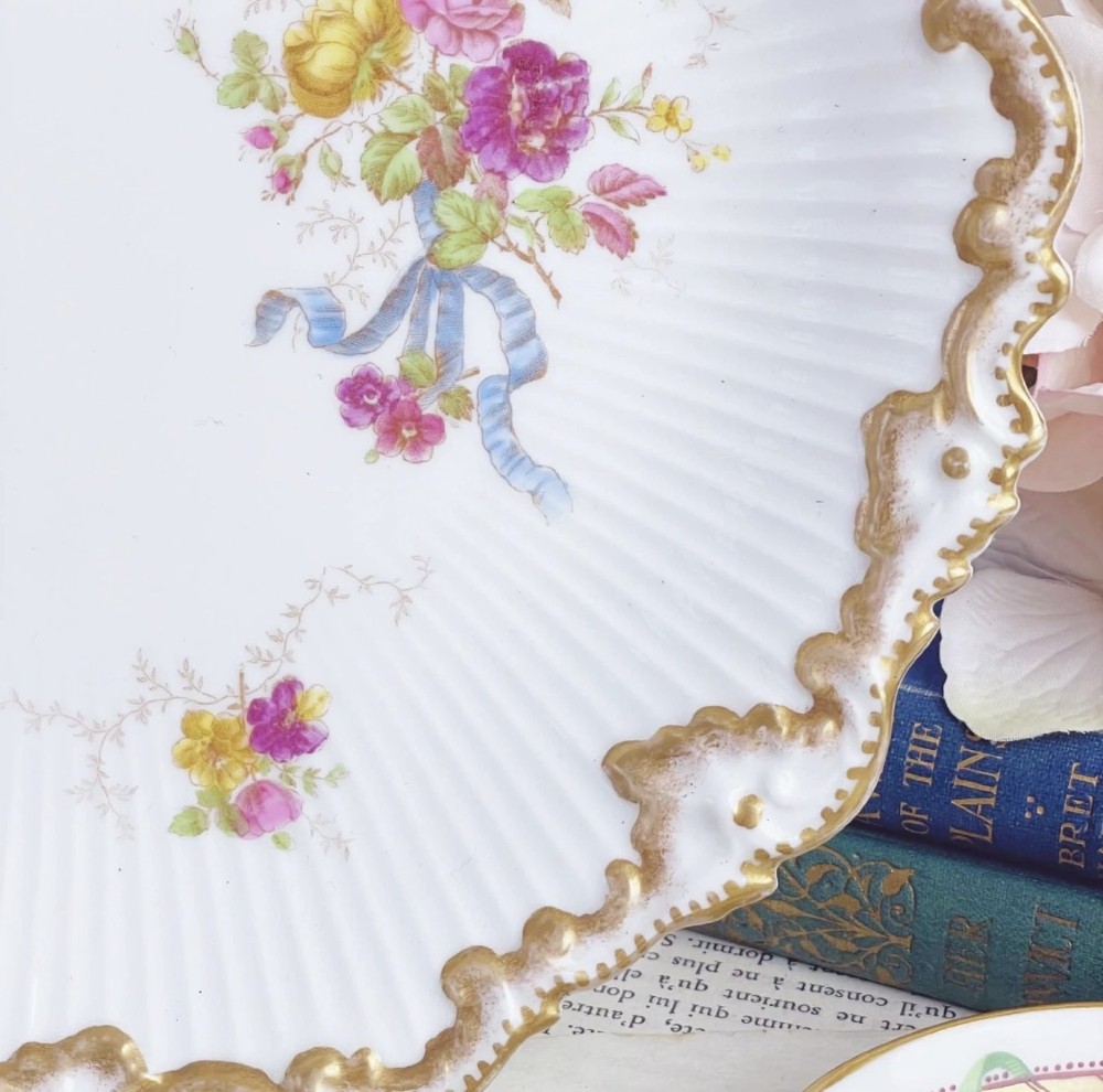 A white circular porcelain plate featuring a gilded scalloped rim, fluted texture, and hand-painted floral sprays adorned with pink and blue ribbons. View III