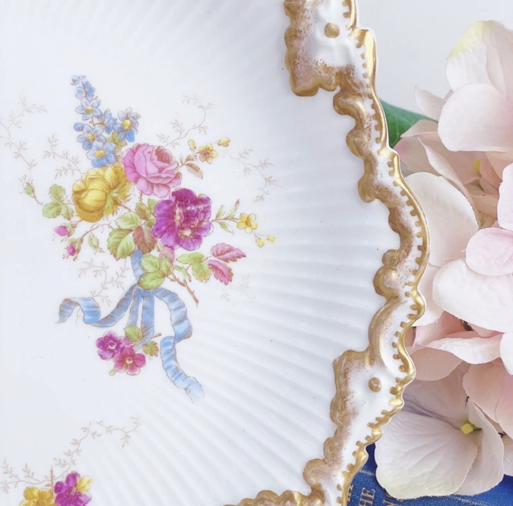 A white circular porcelain plate featuring a gilded scalloped rim, fluted texture, and hand-painted floral sprays adorned with pink and blue ribbons. View IV