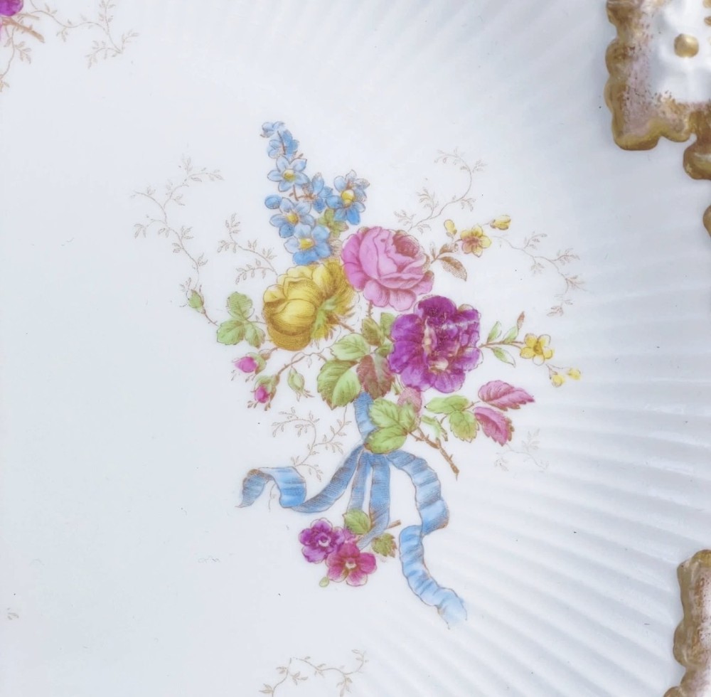 A white circular porcelain plate featuring a gilded scalloped rim, fluted texture, and hand-painted floral sprays adorned with pink and blue ribbons. View V