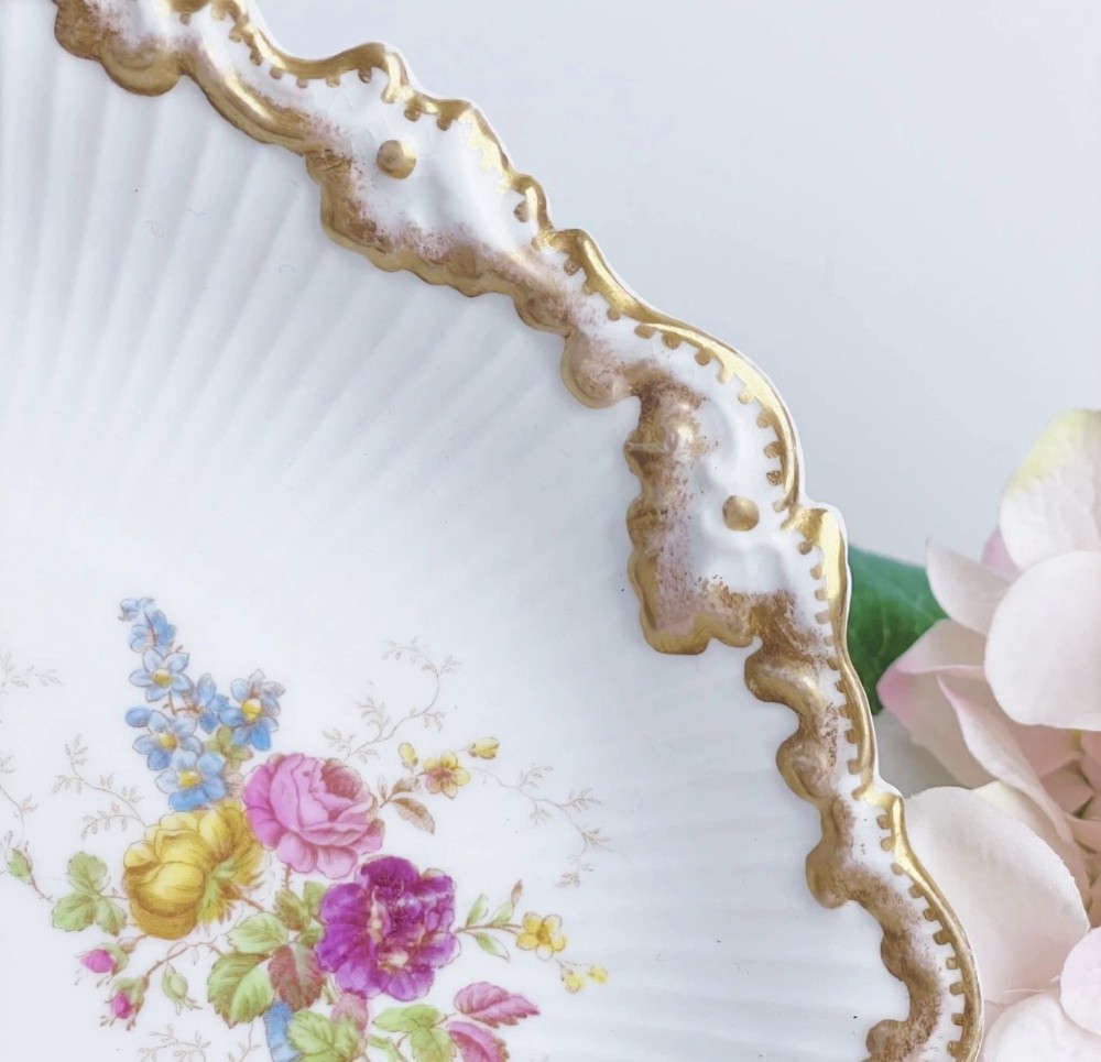 A white circular porcelain plate featuring a gilded scalloped rim, fluted texture, and hand-painted floral sprays adorned with pink and blue ribbons. View VI