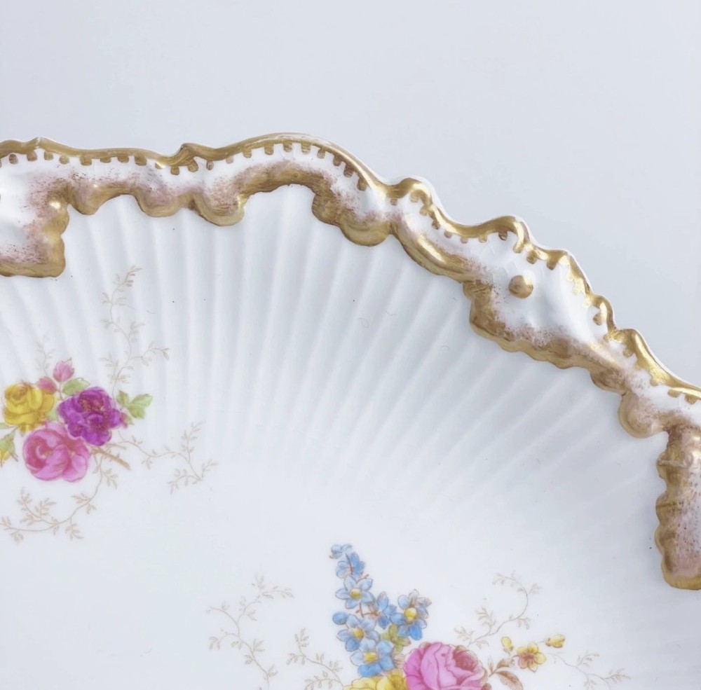 A white circular porcelain plate featuring a gilded scalloped rim, fluted texture, and hand-painted floral sprays adorned with pink and blue ribbons. View VII
