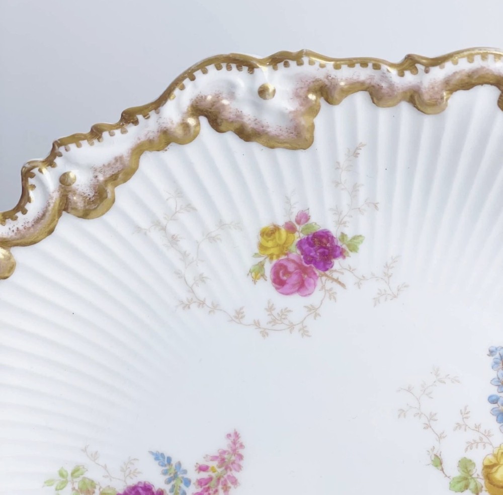 A white circular porcelain plate featuring a gilded scalloped rim, fluted texture, and hand-painted floral sprays adorned with pink and blue ribbons. View VIII