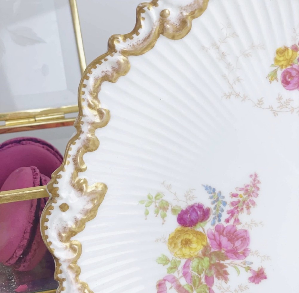 A white circular porcelain plate featuring a gilded scalloped rim, fluted texture, and hand-painted floral sprays adorned with pink and blue ribbons. View IX
