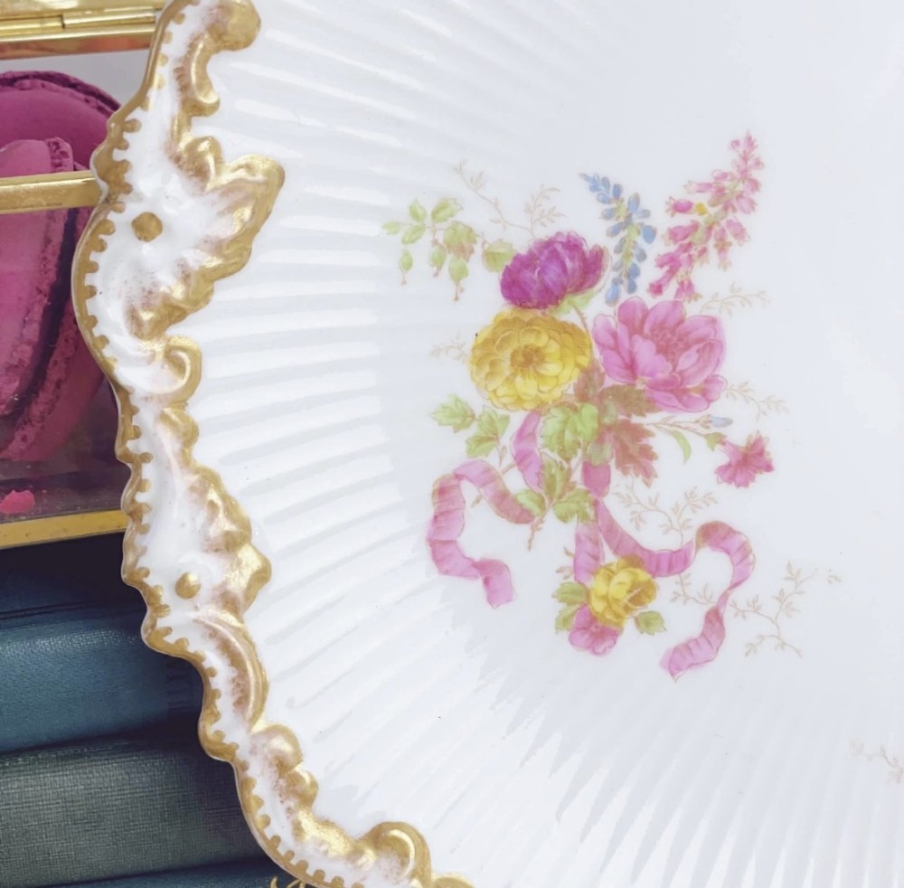 A white circular porcelain plate featuring a gilded scalloped rim, fluted texture, and hand-painted floral sprays adorned with pink and blue ribbons. View X