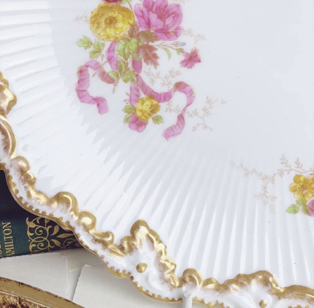 A white circular porcelain plate featuring a gilded scalloped rim, fluted texture, and hand-painted floral sprays adorned with pink and blue ribbons. View XI