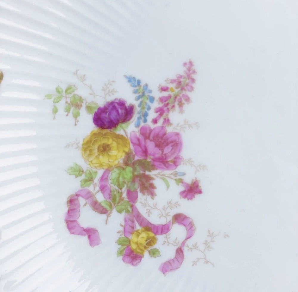 A white circular porcelain plate featuring a gilded scalloped rim, fluted texture, and hand-painted floral sprays adorned with pink and blue ribbons. View XII