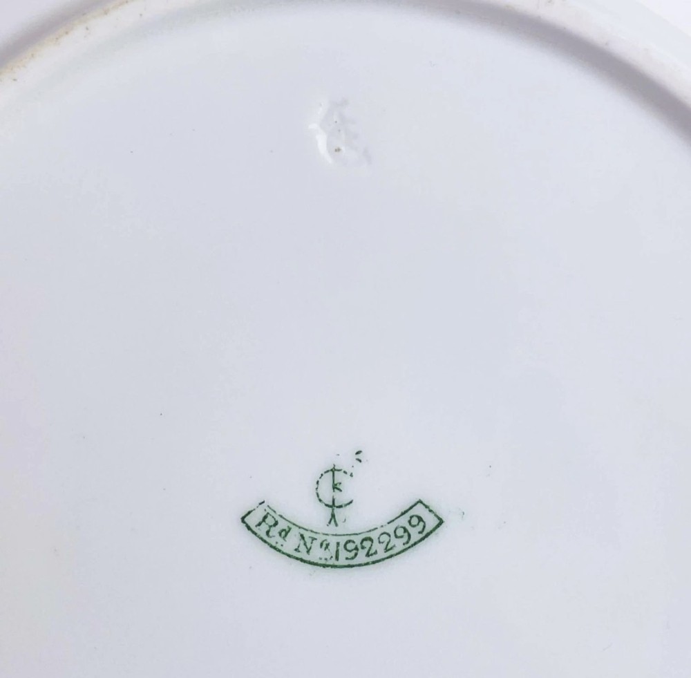 A white circular porcelain plate featuring a gilded scalloped rim, fluted texture, and hand-painted floral sprays adorned with pink and blue ribbons. View XIII