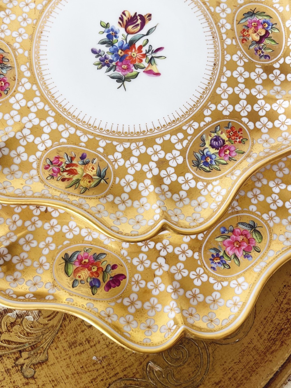 A pair of late Victorian Coalport bone china cabinet plates featuring scalloped rims heavily detailed with gold leaf, white geometric patterns, and central hand-painted floral sprays. View X