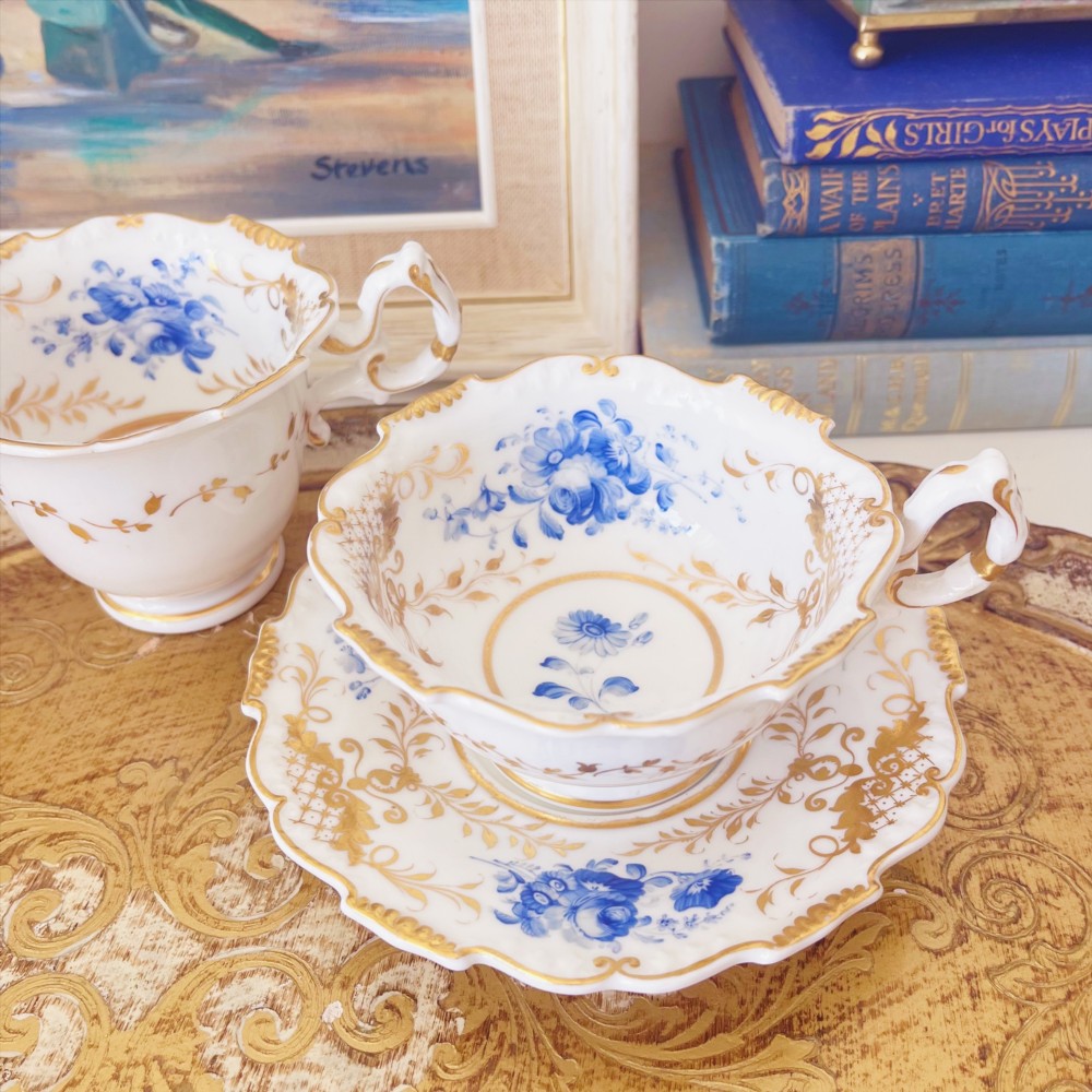 A Regency era porcelain tea trio comprising a teacup, coffee cup, and saucer decorated with blue floral hand-painting and gold leaf gilding.