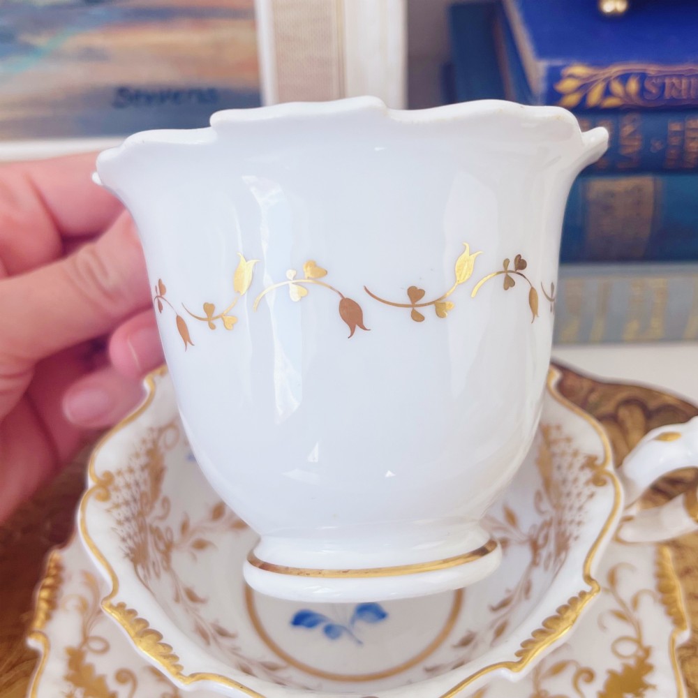 A Regency era porcelain tea trio comprising a teacup, coffee cup, and saucer decorated with blue floral hand-painting and gold leaf gilding. View IV