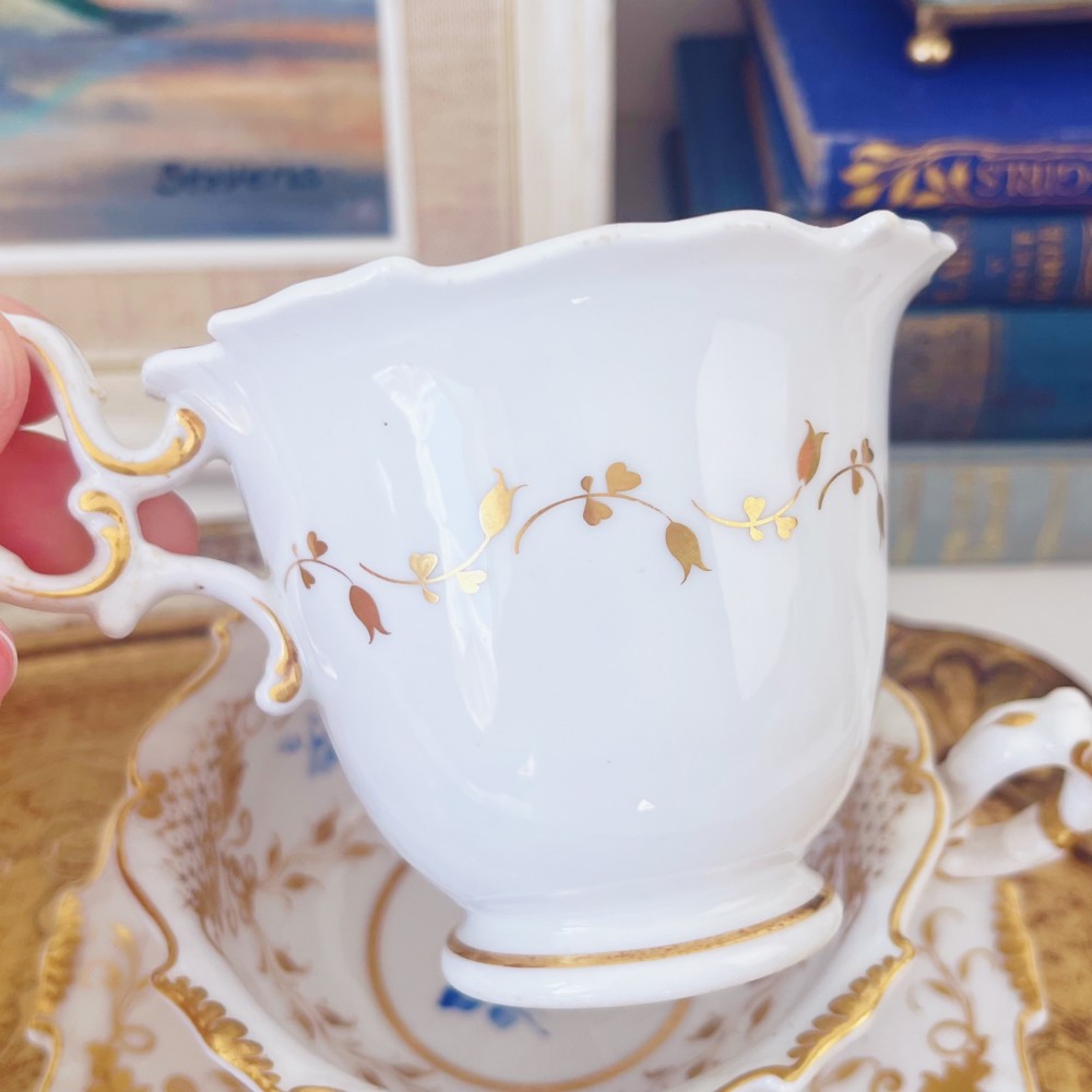 A Regency era porcelain tea trio comprising a teacup, coffee cup, and saucer decorated with blue floral hand-painting and gold leaf gilding. View V