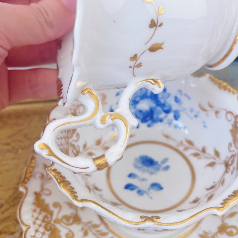 A Regency era porcelain tea trio comprising a teacup, coffee cup, and saucer decorated with blue floral hand-painting and gold leaf gilding. View VI