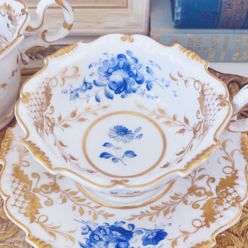 A Regency era porcelain tea trio comprising a teacup, coffee cup, and saucer decorated with blue floral hand-painting and gold leaf gilding. View VII