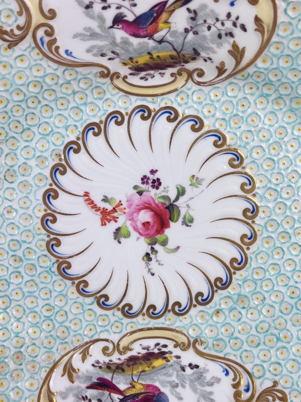 An oval antique porcelain serving dish featuring hand-painted birds in cartouches, a central pink rose, a turquoise pattern ground, and a scalloped gilded rim. View X