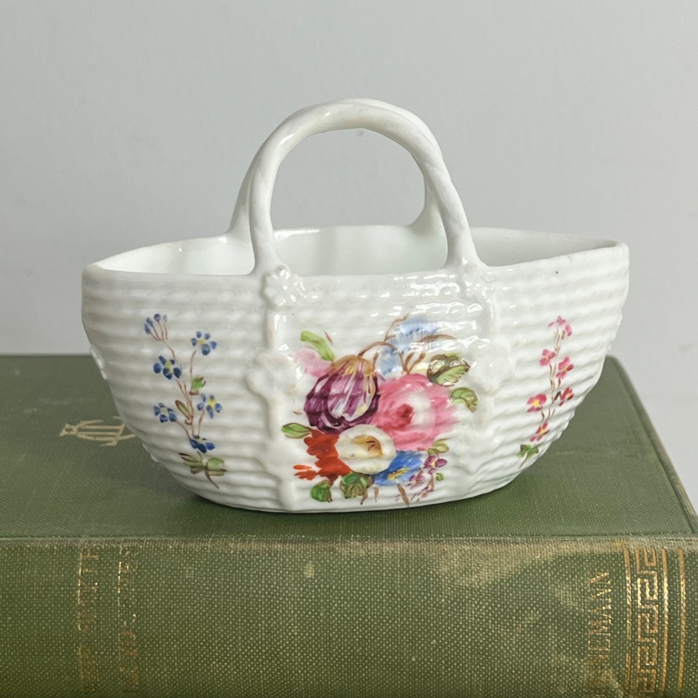 A miniature white porcelain basket featuring a raised basketweave texture, an arched handle, and hand-painted colourful floral motifs on the exterior.