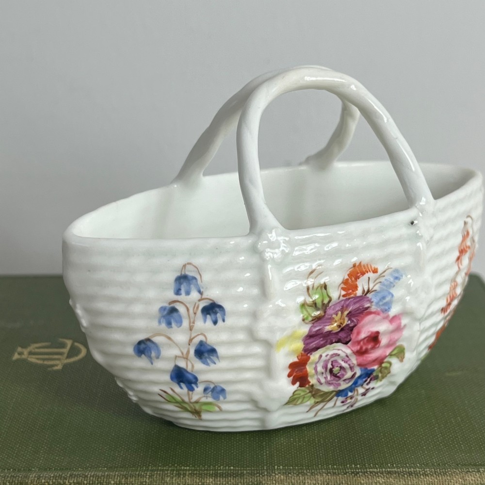 A miniature white porcelain basket featuring a raised basketweave texture, an arched handle, and hand-painted colourful floral motifs on the exterior. View IV