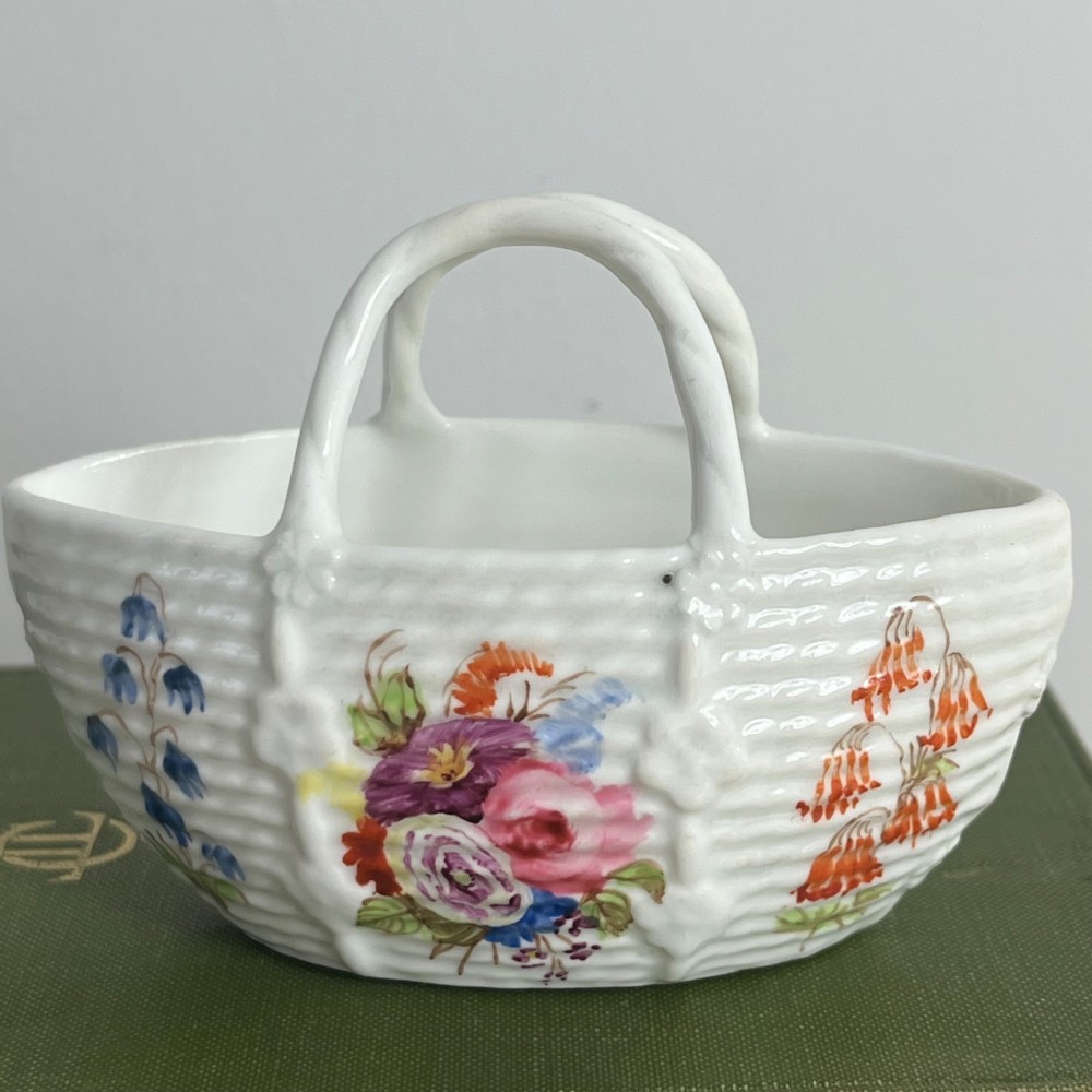 A miniature white porcelain basket featuring a raised basketweave texture, an arched handle, and hand-painted colourful floral motifs on the exterior. View V