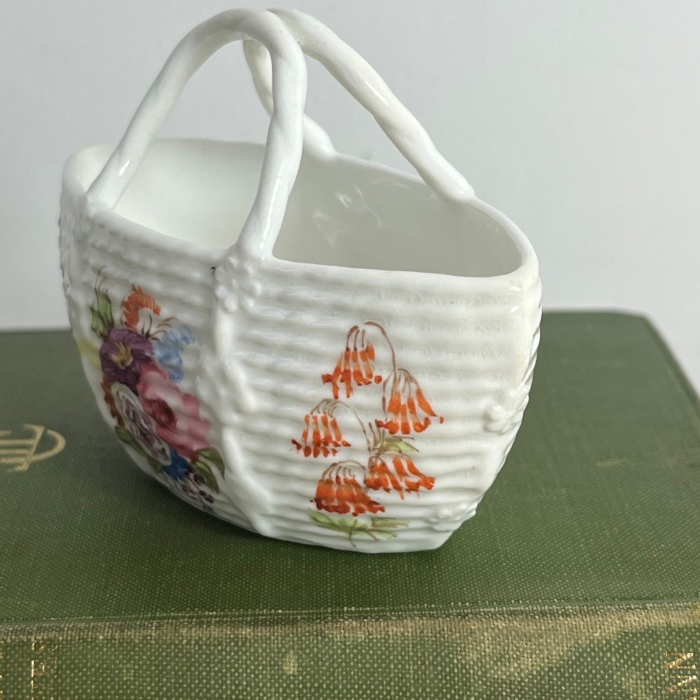 A miniature white porcelain basket featuring a raised basketweave texture, an arched handle, and hand-painted colourful floral motifs on the exterior. View VI