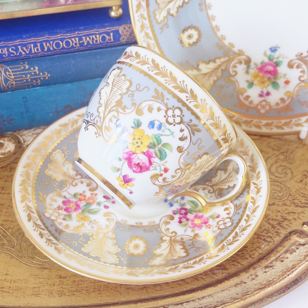 An antique Cauldon bone china teacup trio featuring powder blue borders, intricate gilt scrollwork, and hand-painted floral vignettes on white porcelain. View II