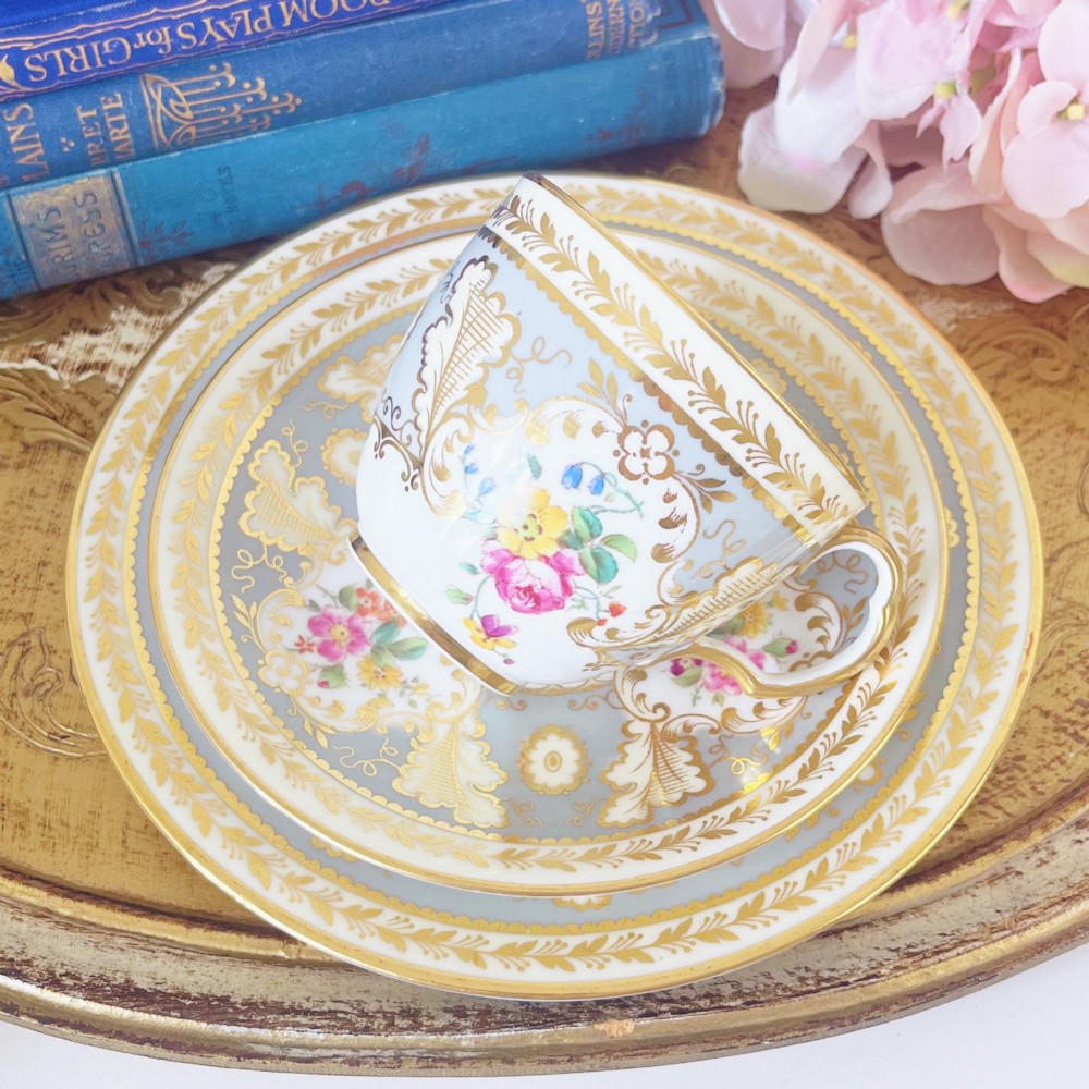 An antique Cauldon bone china teacup trio featuring powder blue borders, intricate gilt scrollwork, and hand-painted floral vignettes on white porcelain. View III