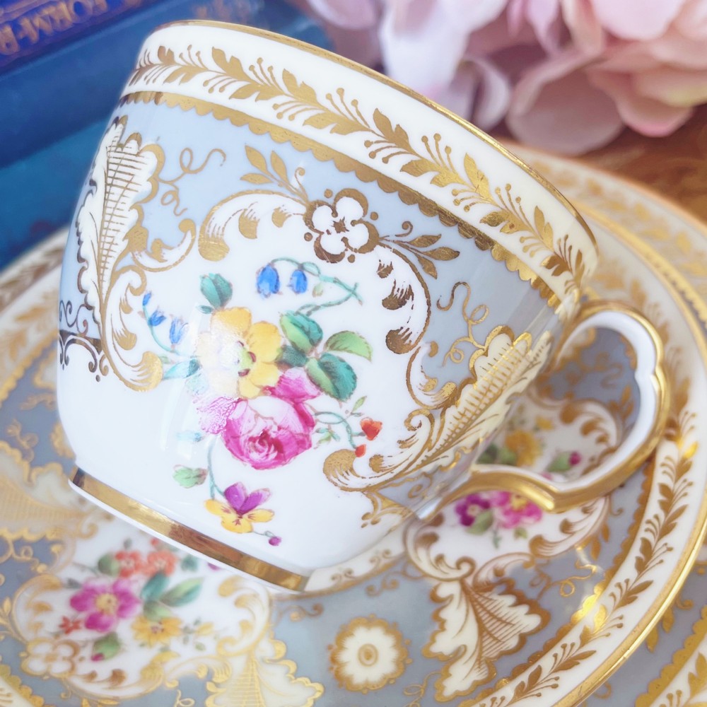 An antique Cauldon bone china teacup trio featuring powder blue borders, intricate gilt scrollwork, and hand-painted floral vignettes on white porcelain. View IV