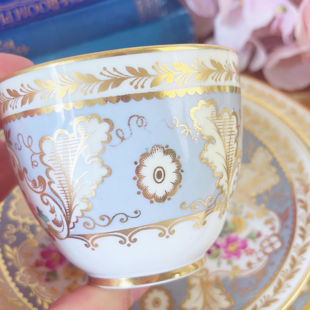 An antique Cauldon bone china teacup trio featuring powder blue borders, intricate gilt scrollwork, and hand-painted floral vignettes on white porcelain. View V