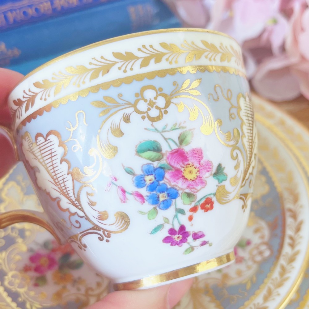 An antique Cauldon bone china teacup trio featuring powder blue borders, intricate gilt scrollwork, and hand-painted floral vignettes on white porcelain. View VI