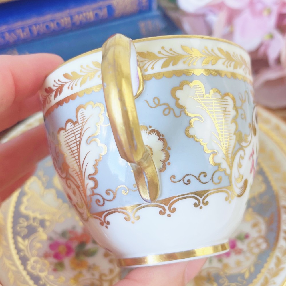 An antique Cauldon bone china teacup trio featuring powder blue borders, intricate gilt scrollwork, and hand-painted floral vignettes on white porcelain. View VII