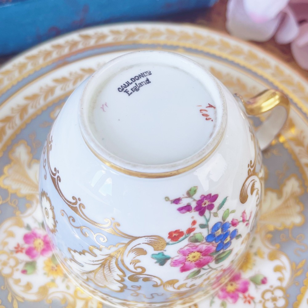 An antique Cauldon bone china teacup trio featuring powder blue borders, intricate gilt scrollwork, and hand-painted floral vignettes on white porcelain. View X