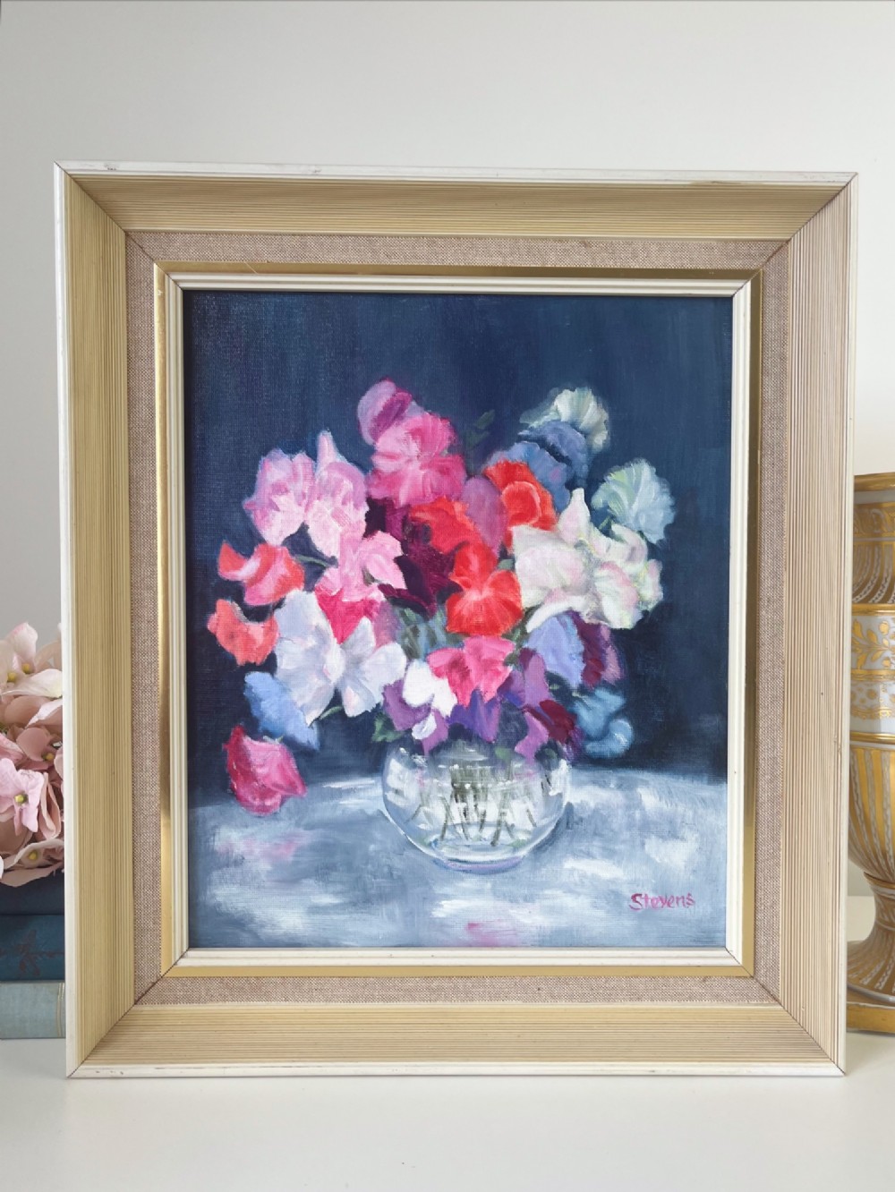 A framed oil painting depicting a colourful bouquet of pink, red, and purple sweet peas in a clear glass vase set against a dark blue background.