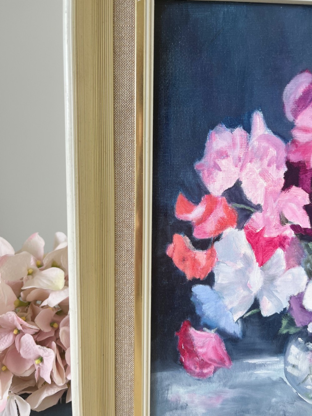 A framed oil painting depicting a colourful bouquet of pink, red, and purple sweet peas in a clear glass vase set against a dark blue background. View VI