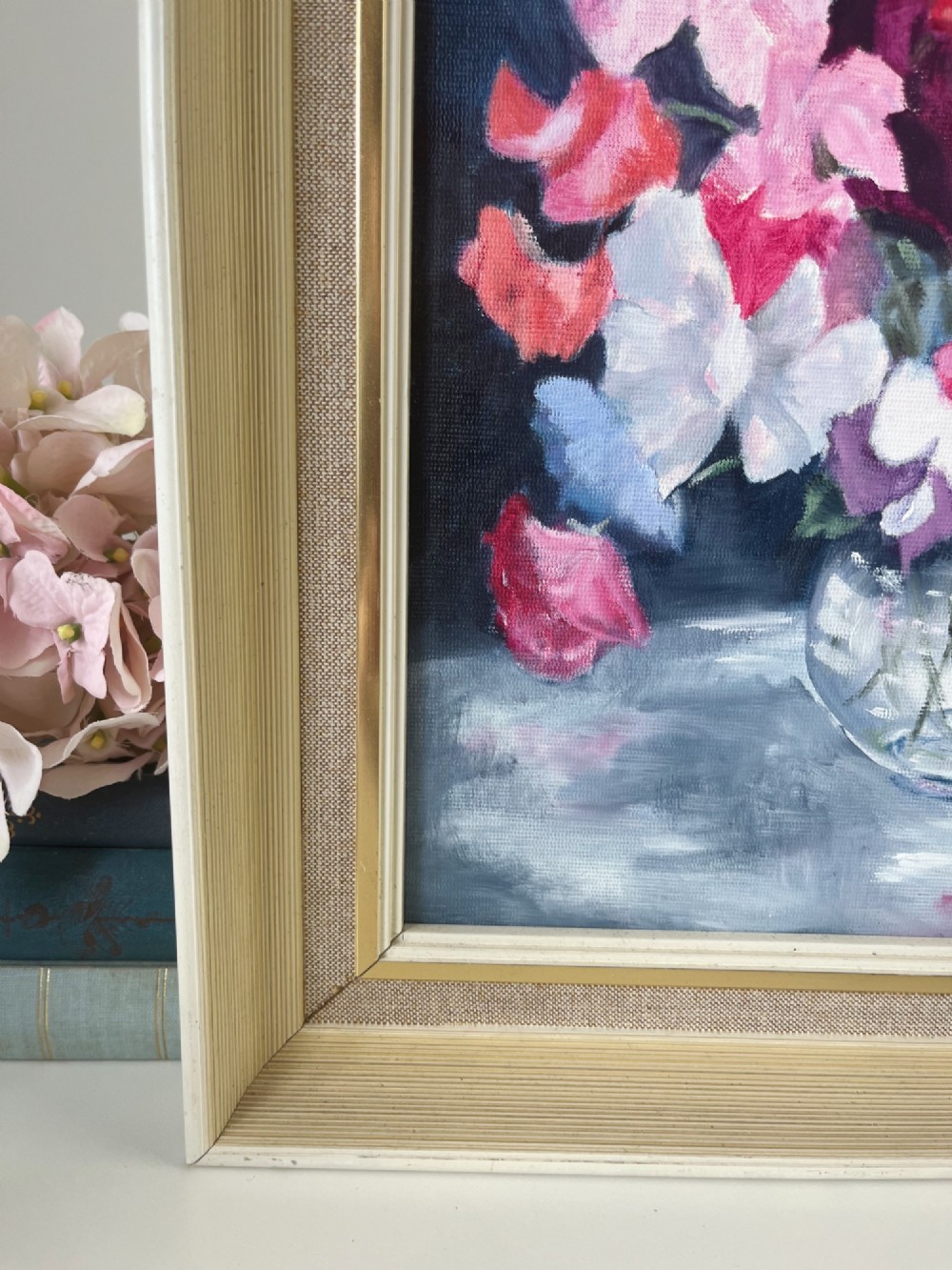 A framed oil painting depicting a colourful bouquet of pink, red, and purple sweet peas in a clear glass vase set against a dark blue background. View VII