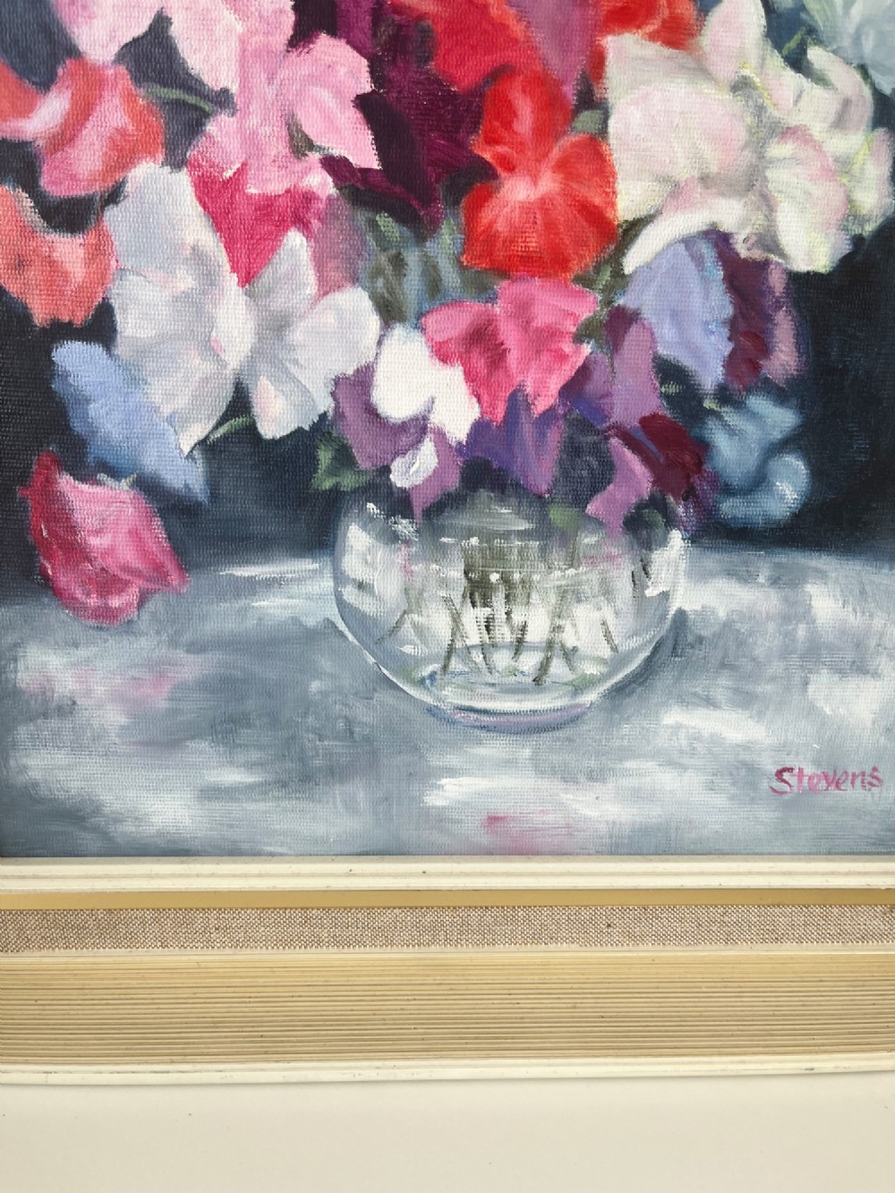 A framed oil painting depicting a colourful bouquet of pink, red, and purple sweet peas in a clear glass vase set against a dark blue background. View VIII