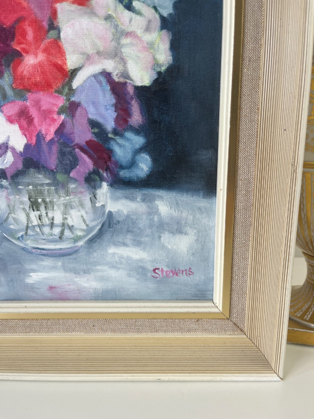 A framed oil painting depicting a colourful bouquet of pink, red, and purple sweet peas in a clear glass vase set against a dark blue background. View IX