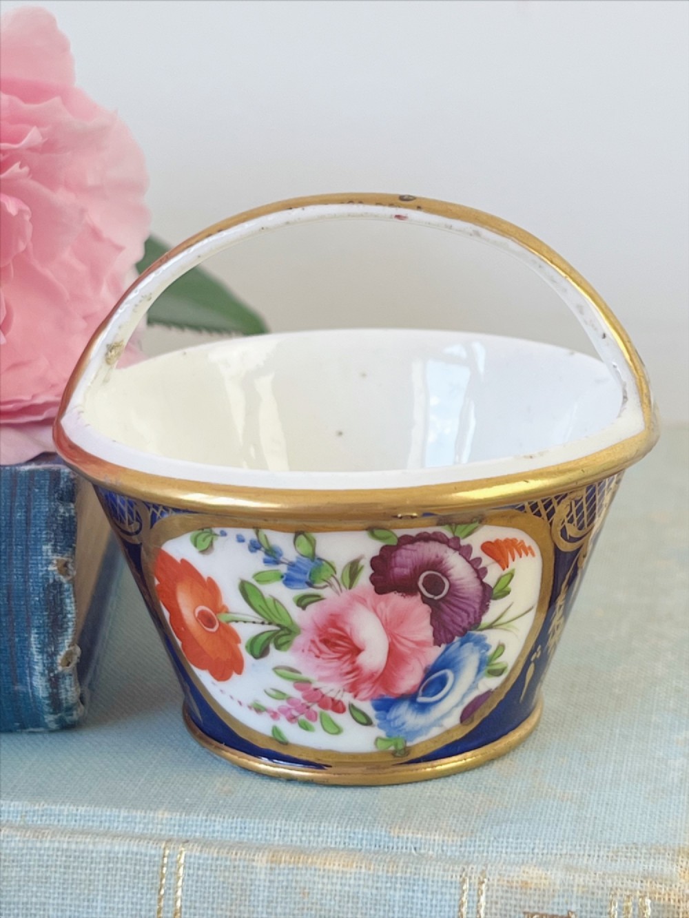 A miniature Regency Coalport porcelain basket featuring an overhead gilt handle, cobalt blue exterior casing with a hand-painted floral reserve, and a plain white interior.